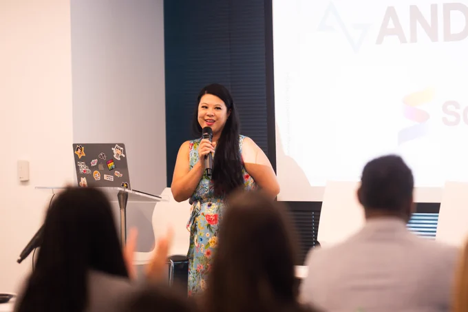 Alt text: A speaker stands at the front of a conference room holding a microphone and addressing an audience. She wears a blue floral dress and stands beside a clear podium with a sticker-covered laptop. Attendees are visible in the foreground, slightly out of focus, while a presentation screen with event branding is displayed behind her.