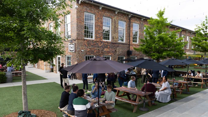People are seated at picnic tables with umbrellas in a grassy outdoor area next to a brick building. More people are gathered nearby, socializing. The setting is a casual gathering with trees and string lights enhancing the ambiance.