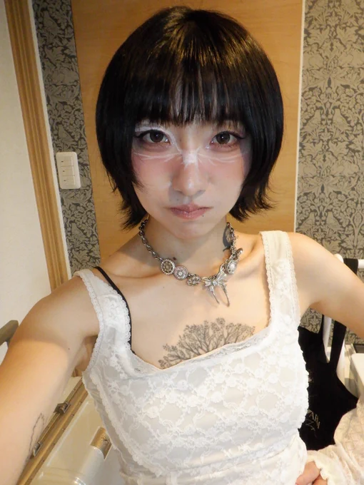 A close-up selfie showing white face paint forming a starburst pattern across the nose and under the eyes, a silver ornate necklace, a tree tattoo on the chest, and a white lace top. The background features patterned wallpaper.

鼻の付け根から目の下にかけて白いフェイスペイントで星形模様が描かれた人物のクローズアップセルフィー。シルバーの装飾的なネックレス、胸元の木のタトゥー、白いレーストップス。背景には柄物の壁紙。