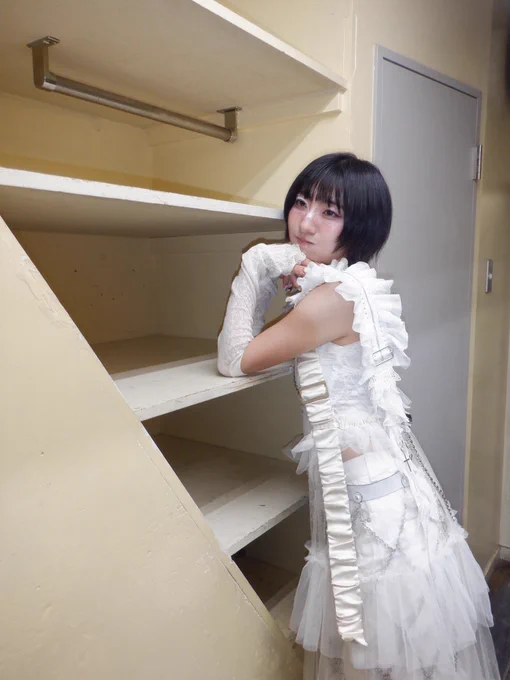 A person in an elaborate white layered outfit with lace gloves, ruffles, buckles, chain details, and a tulle skirt leans against an empty wooden shelf unit, looking over their shoulder with a calm expression.

レースグローブ、フリル、バックル、チェーン装飾、チュールスカートなど装飾豊かな白いコーデを身にまとった人物が、空の木製棚に寄りかかり、肩越しに静かな表情でカメラを見ている。