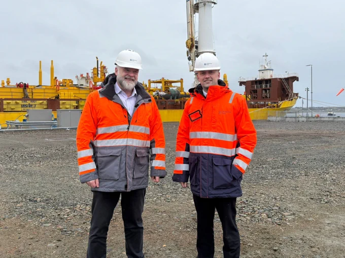 Transport Secretary Stephen Flynn poses for a photo with Alex MacLeod, Chief Executive of Stornoway Port Authority. They are wearing hi-vis clothing and hard hats and there is a freight tanker behind them.