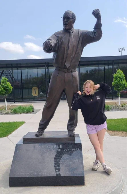 A bronze statue of a man in a dynamic, triumphant pose stands on a pedestal outdoors. Beside it, a smiling person in a black hoodie and purple shorts playfully mimics the statue's pose. The background features a modern glass building and lush greenery.