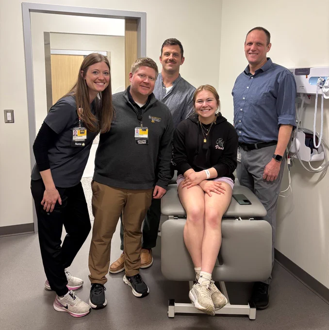 A group of four smiling adults stands next to a young woman sitting on a medical examination table. The woman is wearing a black hoodie and shorts, with a hospital wristband. The others are dressed in professional attire, and one wears a nurse's uniform. The setting appears to be a clinic room with medical equipment on the wall.