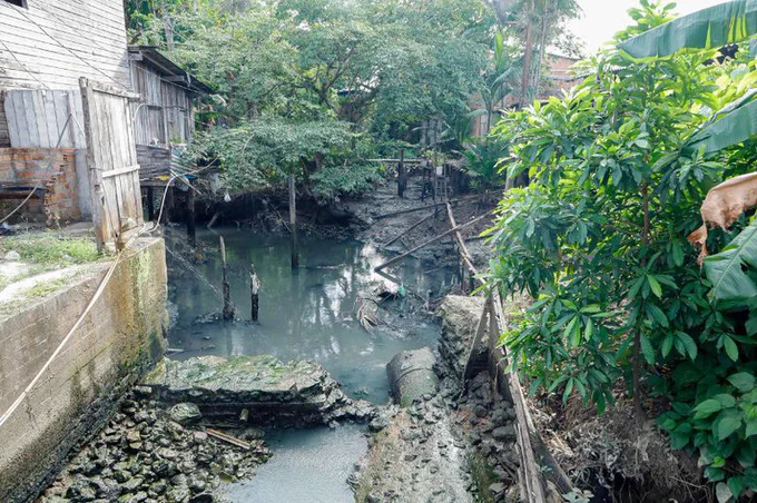 Vista de um canal com água escura e detritos, cercado por vegetação densa e casas rústicas de madeira nas laterais. Foto: João Gomes/Comus/Prefeitura de Belém.