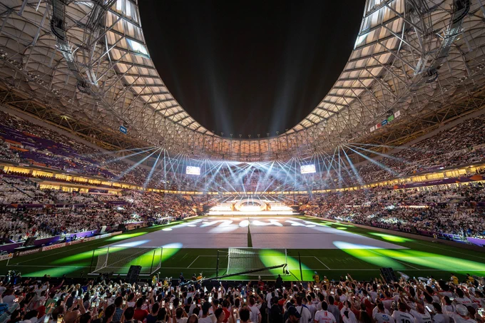 A brightly lit stadium packed with spectators, featuring an expansive field. Spotlights illuminate the scene, and large banners with logos and advertisements are visible around the stadium.