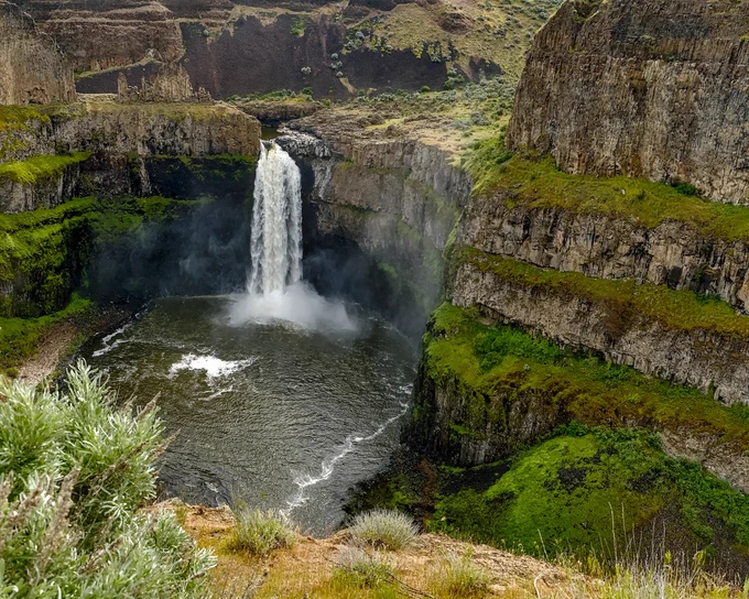 A wide view of Palouse Falls dropping from a basalt cliff into a deep circular pool below. Steep canyon walls surround the waterfall, with layers of dark volcanic rock, tan grasses, and bright green spring growth covering parts of the cliffs. Mist rises from the base of the falls, and the river curves through the pool below. The photo feels dramatic, rugged, and full of natural power.