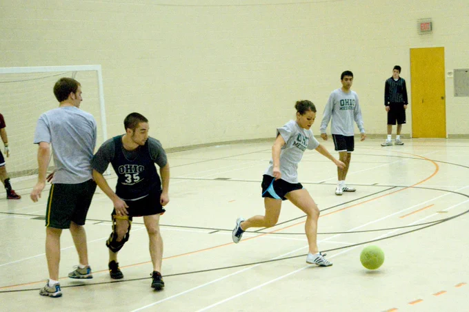 A student runs down the gymnasium court, dribbling a bright green indoor soccer ball. Other players are actively moving around the court to defend and follow the play.