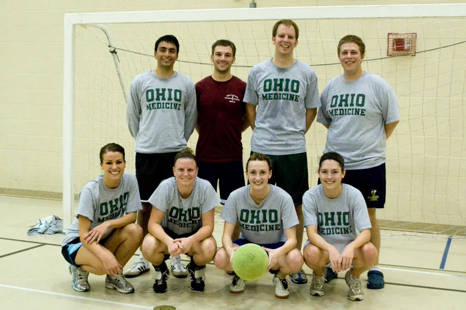 A smiling group of eight Heritage College students poses together for a team photo in front of an indoor soccer net, with one student in the front holding the green soccer ball.