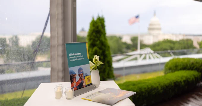 A table on a terrace overlooks the U.S. Capitol building in the distance. On the table, there is a small sign and pamphlet. An American flag is visible in the background.