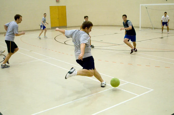 A student winds up to kick a bright green indoor soccer ball across a gymnasium court. Several other students can be seen running and watching the play in the background.