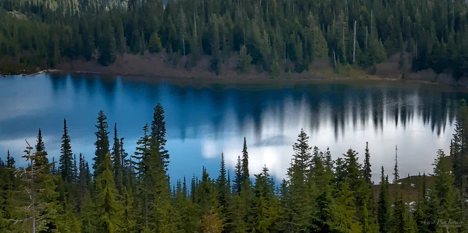 A landscape photo looking across a calm mountain lake surrounded by evergreen forest. The blue water reflects dark tree shapes and soft white clouds, creating a mirrored band across the center of the image. Evergreen trees fill the foreground and distant shoreline, with subtle patches of snow visible near the lower edge. The scene feels quiet, layered, and still. The photo makes me feel peaceful and drawn into the calm reflection of the mountain landscape.