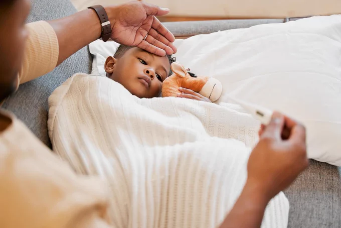 Child lying on a couch covered with a white blanket, holding a stuffed animal while parent checks their temperature with a thermometer.