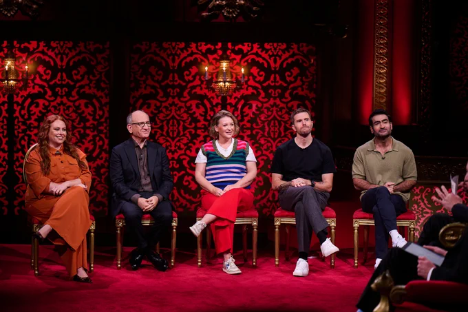 Five contestants sit on red velvet chairs in a richly decorated studio with red patterned walls and chandelier lighting during a Taskmaster recording. From left to right are Amy Gledhill, Armando Iannucci, Joanna Page, Joel Dommett and Kumail Nanjiani. They face toward the host, who is partially visible in the foreground, while the audience area is out of frame. The set features ornate gold detailing and a red carpet, creating a theatrical atmosphere.