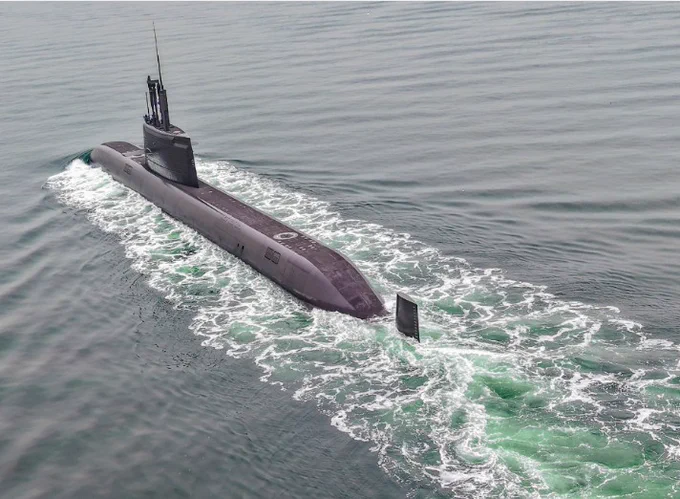 A military submarine cruising on the ocean surface, leaving a trail of white foam in its wake.