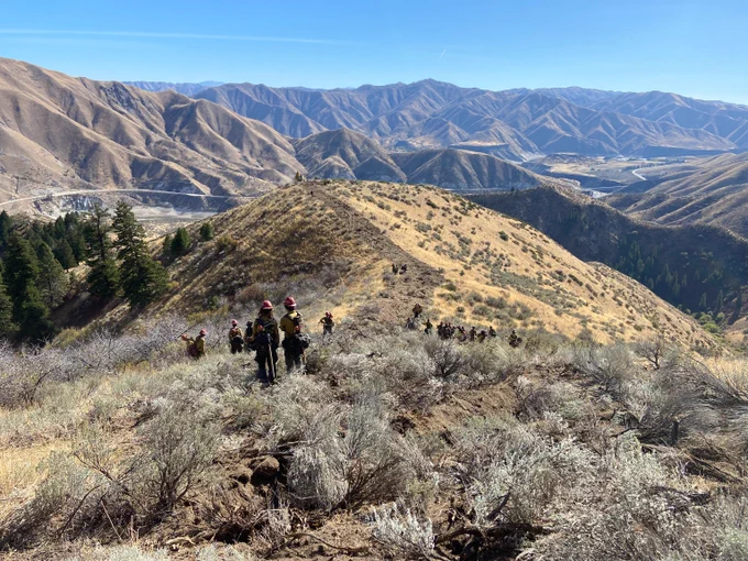 Group of wildland firefighters trekking along a dry, shrub-covered ridge with expansive mountain ranges in the background under a clear blue sky.