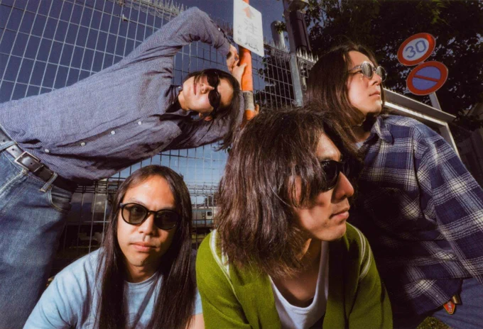 Band photo of the four members of DYGL posing in front of a camera, but most are not looking into the lens and two of them are even looking away. The musicians are dressed casually and all wearing sun glasses. They are posing in front of a gate and a train is visible in the background. Photography: Earl Standerford