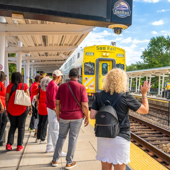 A group of people standing on a SunRail station platform watching as a SunRail train approaches under a clear sky.