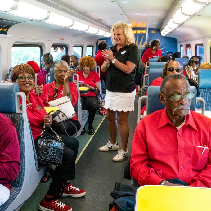 A group of people wearing red shirts are seated inside a SunRail train while the group ride organizer stands smiling with them. 