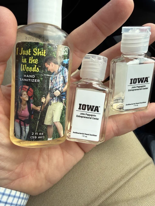 A photo of Rob Sand holding three small bottles of hand sanitizer. One bottle is labeled "I Just Shit in the Woods Hand Sanitizer" with an image of two people hiking. The other two bottles are labeled "IOWA John Pappajohn Entrepreneurial Center."