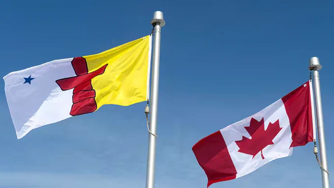 Alt text:

Two flags fly side by side on tall flagpoles against a clear blue sky. On the left is the flag of Nunavut, featuring a yellow and white background divided vertically, a red inuksuk at the centre, and a blue star near the upper left corner representing the North Star (Niqirtsuituq). On the right is the Canadian flag, displaying a red maple leaf between two red vertical bands. Both flags are billowing in the wind, symbolizing the relationship between Nunavut and Canada and the recognition of Inuit culture and self-governance within the Canadian federation.