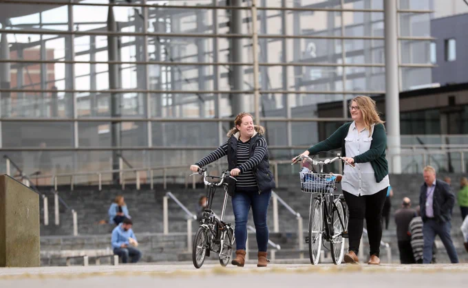 Two women walking bicycles in front of the Senedd.  Dwy fenyw yn cerdded beiciau o flaen y Senedd.