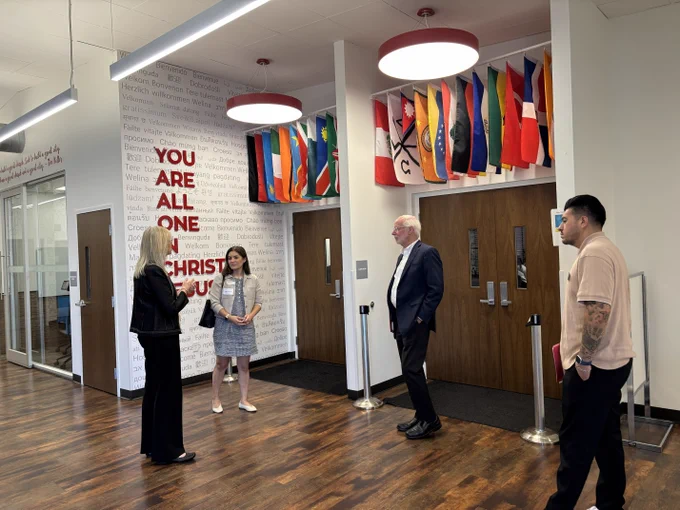 Four people convening in a hallway decorated with international flags and a wall sign reading 'You Are All One in Christ Jesus.'