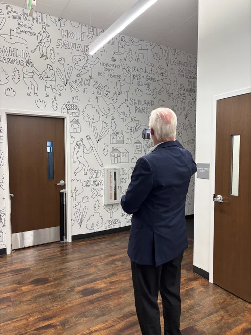 JB Holston stands in a hallway taking a photo of a white wall with black line art illustrations and text.