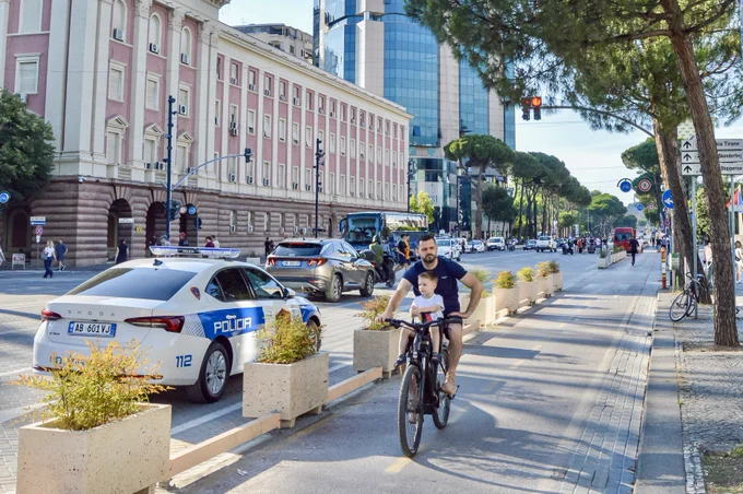 People of all ages and abilities ride bicycles along protected infrastructure lined with buildings, trees, lighting, and signage in.central Tirana.