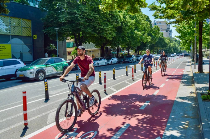 People of all ages and abilities ride bicycles along protected infrastructure lined with buildings, trees, lighting, and signage in.central Tirana.