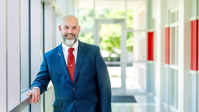 Aaron Kiess in a navy blue suit and red tie standing confidently in a bright modern hallway with large windows.