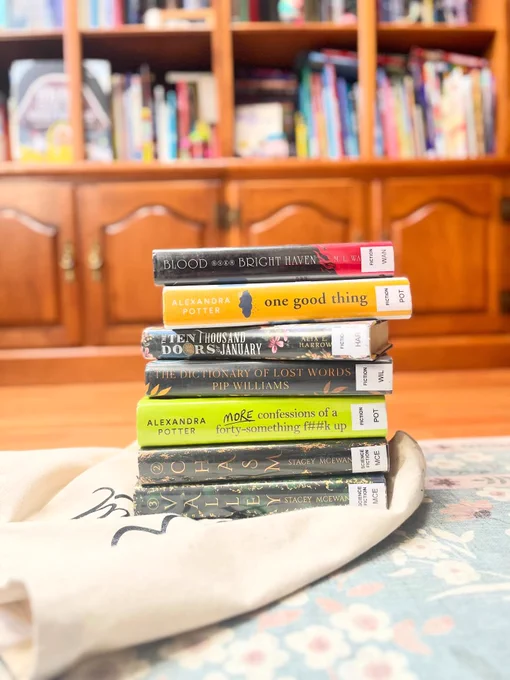 Photo of a stack of eight books on a floral patterned surface in front of a wooden bookshelf filled with more books. 