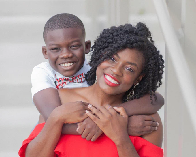 African American boy and his mother looking at the camera and smiling. Mom is wearing a red dress. Boy is wearing a white shirt with a black, red, and white bow tie.