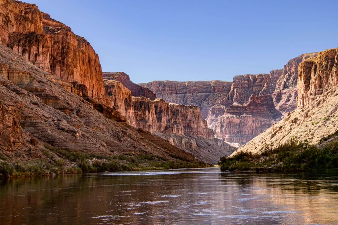 Wide view of a calm river flowing through towering red rock canyon walls under a clear blue sky.
