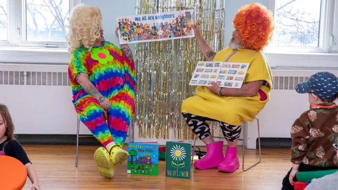 Two drag performers, dressed in colourful outfits, conduct storytime in the library. They are sitting in chairs in front of a wall decorated with shiny hanging tassels. They hold a picture book between them, the pages open to an illustration of a crowd of diverse people. The large text in the book reads "We are all neighbours.