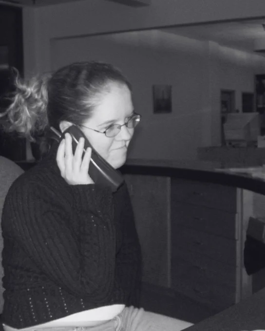 Woman with a messy bun wearing a dark sweater is seated indoors, holding a phone to their ear.