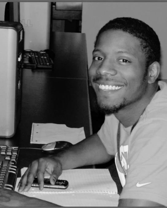 Man with a smile seated at a desk using a computer keyboard with a notebook and phone nearby in a workspace.