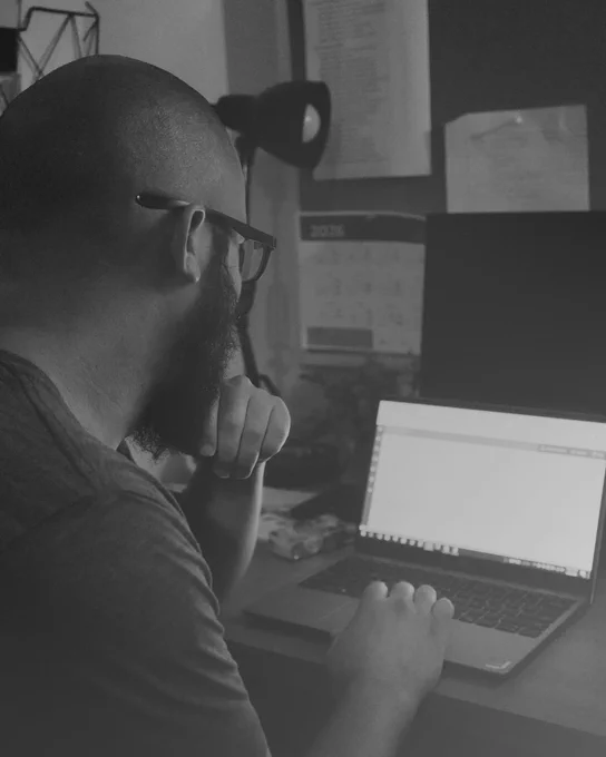 Man with glasses and beard working thoughtfully on a laptop at a desk in a dimly lit room.