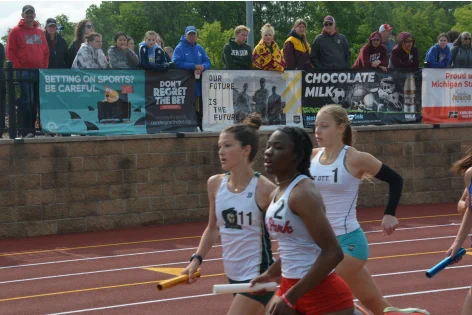 Three female athletes sprinting on a track relay race, each holding a baton, with spectators watching behind a barrier displaying various banners.