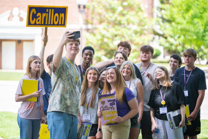 A group of people posing for a selfie outside. One person holds a sign that says "Carillon." Others hold folders and signs, including one with "Golden Eagle." They are in a campus-like setting with trees and a building in the background.