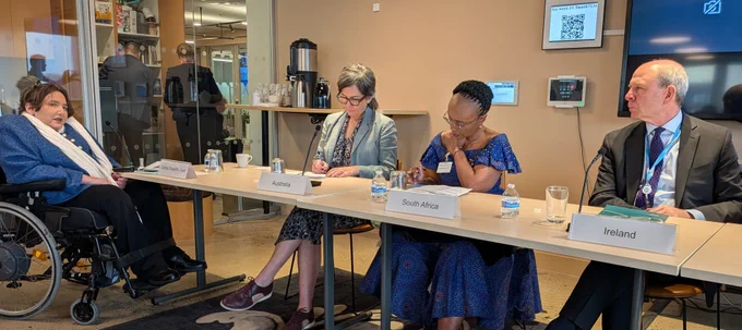 A small meeting room panel: four participants sit at a table with country nameplates (“Australia,” “South Africa,” “Ireland”) and microphones. One participant uses a wheelchair. Papers, notebooks, and water glasses are on the table. Behind glass, a kitchenette with cups and coffee urns is visible.