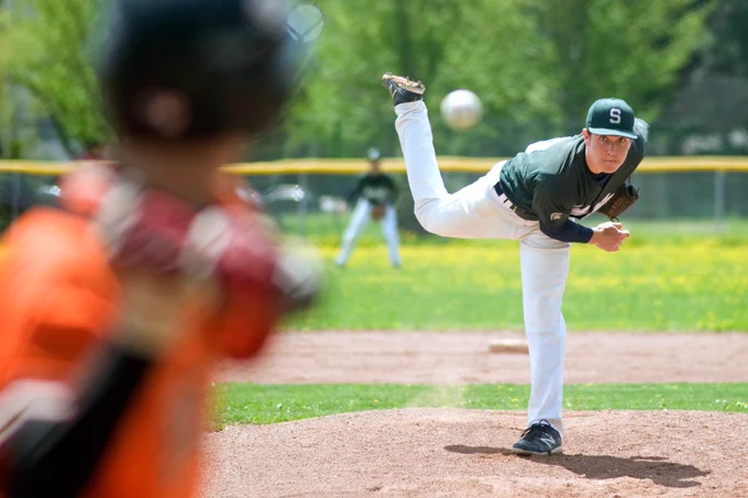 A baseball pitcher throws a pitch from the mound, with the ball mid-air on the way to the batter. The camera focuses on the pitcher in the follow-through motion.