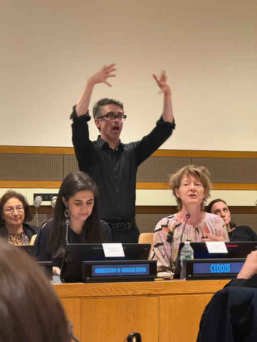A conference room panel is shown. Several people sit at a long desk with microphones and nameplates; visible text includes “Organization of American States” and “CEDDIS.” One person in black stands behind the panel, raising both hands in expressive gestures, likely interpreting or emphasizing speech. Others look down at notes or forward. Background shows a plain wall and wood paneling.
