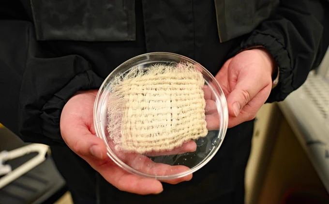 A person in a black jacket holds a clear petri dish with both hands. Inside the dish is a rectangular piece of light-beige, coarsely woven fabric, heavily frayed on one side, and submerged in a small amount of liquid.