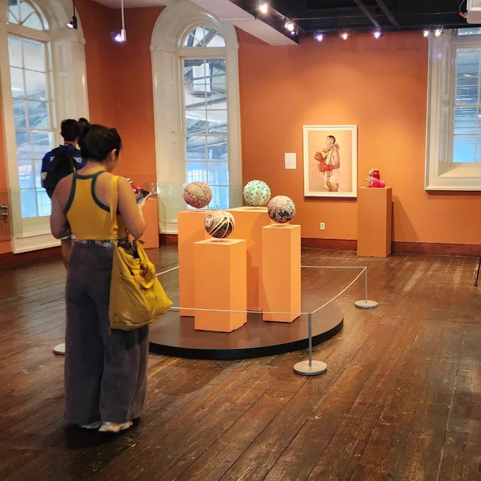 Visitors observe colorful spherical art pieces displayed on orange pedestals in a warmly lit gallery with wooden floors and large windows.