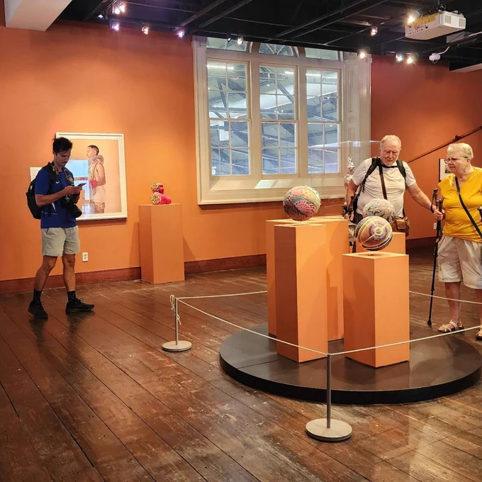 Visitors observe colorful spherical sculptures displayed on orange pedestals in a warmly lit art gallery with wooden floors and large windows.