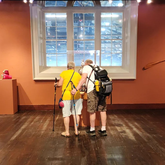 Two elderly people with walking sticks closely examining an exhibit in a warmly lit museum room with large windows.