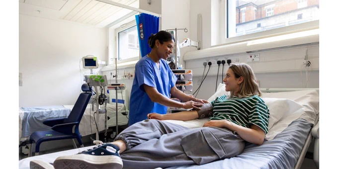 Healthcare worker checks a patient’s blood pressure while the patient rests in a hospital bed, with medical equipment visible in a bright clinical room