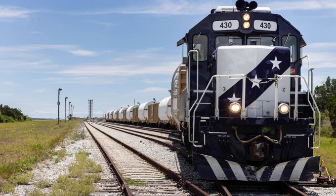A gray and black locomotive, marked with three stars on its front, pulls a long line of white rail cars along tracks surrounded by gravel. A bright blue sky provides a clear backdrop. Credit: NASA/Cory S. Huston