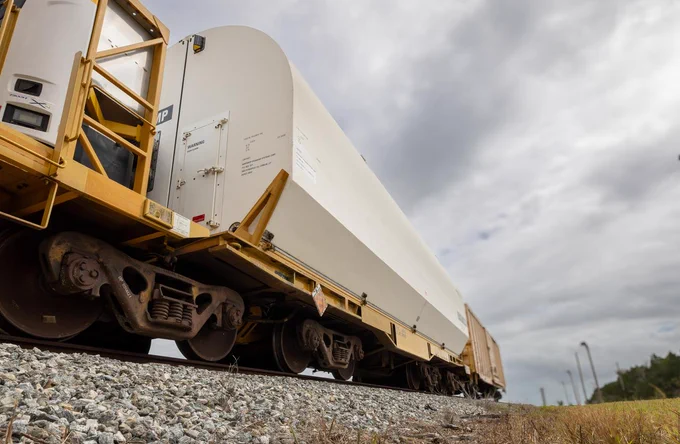 Taken from a low, ground-level perspective, the image shows gravel and grass in the foreground as a white train car passes overhead. The camera angle looks upward, emphasizing the train's size against a backdrop of gray, overcast clouds. Credit: NASA/Frank Michaux