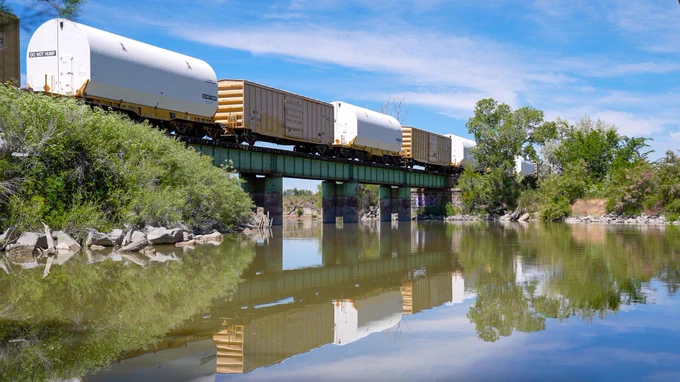 A train crosses a bridge over calm water, its reflection mirrored below. Lush green vegetation frames the scene, and clear blue skies on a sunny day create a bright, picturesque landscape. Credit: NASA/Brandon Hancock